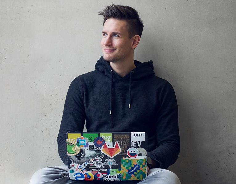 Simon lacht freundlich und sitzt mit seinem MacBook auf dem Boden. Anscheinend kann er sich keinen Schreibtisch leisten.