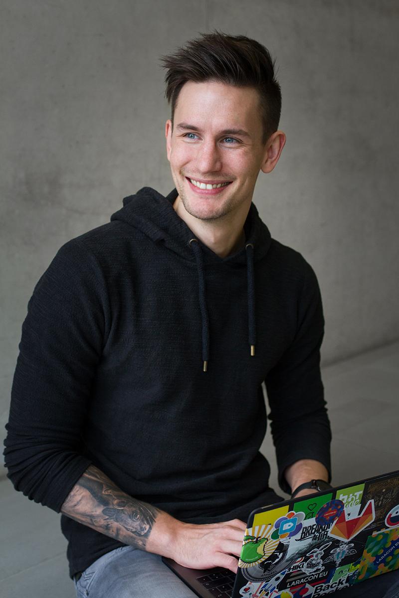 Simon lacht freundlich und sitzt mit seinem MacBook auf dem Boden. Anscheinend kann er sich keinen Schreibtisch leisten.